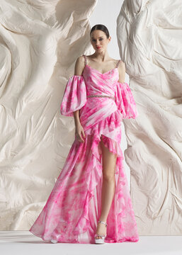 Full length shot of elegant woman wearing luxury pink floral watercolor printed organza gown with puff sleeves and ruffled hem posing gracefully against sculptural white studio wall backdrop