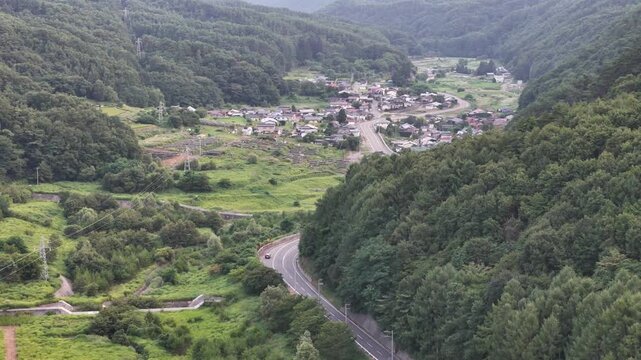 Green valley cradles quiet village nestled between forested hills. Winding road cuts through lush meadows, inviting exploration. Tiny houses cluster near open fields, blending nature