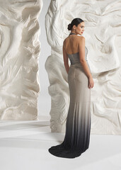 Rear back view of elegant woman wearing luxury silver ombre metallic gradient ombre jersey dress with knot drapery and open back posing gracefully against sculptural white studio wall © Cumhur