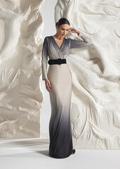 Full length shot of elegant woman wearing luxury silver ombre metallic gradient pleated gown with black waist belt and deep V posing gracefully against sculptural white studio wall backdrop © Cumhur