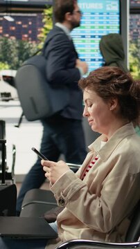 Vertical frame: Smiling woman texts on a smartphone in a bus station while waiting for an intercity coach connection, chatting with friends online on her mobile phone during the wait before departure.