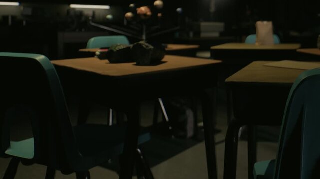 Several empty tables and chairs are arranged in a dark room. The light source is minimal, creating shadows around the furniture. It appears to be evening inside a building.