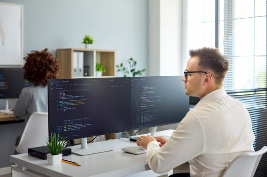 Programmer developer coding software on monitors in office. Focused male engineer at dual screens, writing clean code in bright workspace. Clear modern software development teamwork.