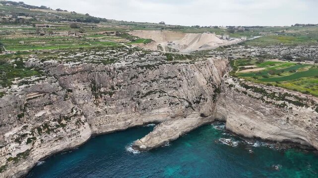 limestone sea cave cove xaqqa cliffs malta aerial drone view stone quarry pit terraced farmland clifftop rugged cliff face waves breaking mediterranean coastline overcast sky