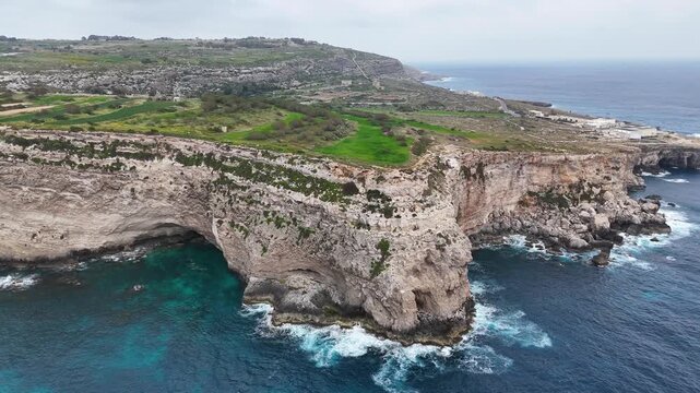 broad sea cave undercuts limestone headland xaqqa cliffs malta aerial drone view hillside village green farmland turquoise water waves crashing coastal cliffs overcast sky