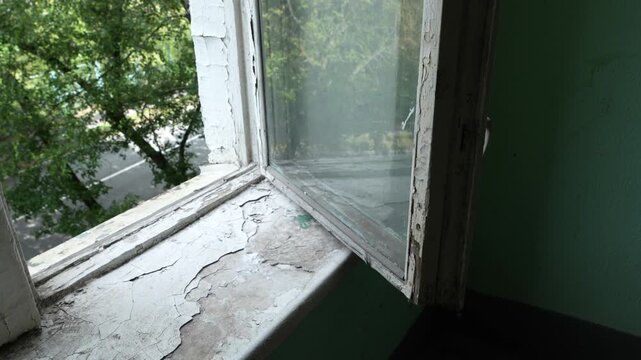 Old residential stairwell window with peeling paint, cracked windowsill and deteriorating surfaces, illustrating aging housing infrastructure, property maintenance and building renovation needs