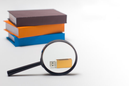 Magnifying glass focusing on a gold USB flash drive with a stack of books blurred in the background, symbolizing digital data search, information storage, and modern education technology.