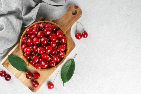 Fresh ripe sweet cherries in rustic wooden bowl with green leaves on light textured background. Top view flat lay with copy space. Juicy organic summer fruit, red berries, healthy seasonal food