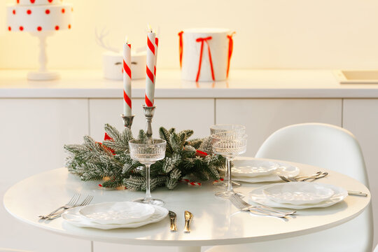 Elegant Christmas table setting with red and white striped candles, evergreen branches, red bows, crystal glasses and white plates in a bright dining room on window background.