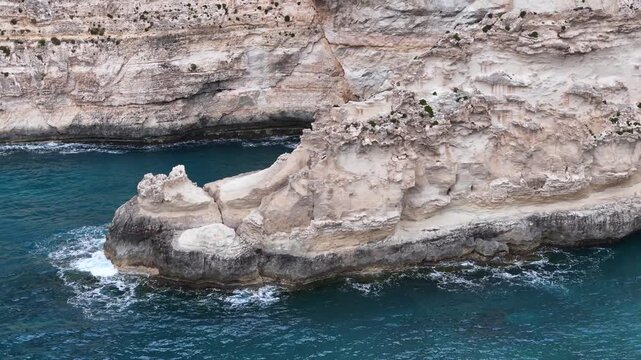 limestone sea cliff promontory with pale cream rock divides a turquoise cove an undercut sea cave shadows its base whitewater surf breaks on rugged malta coastline aerial drone flyover