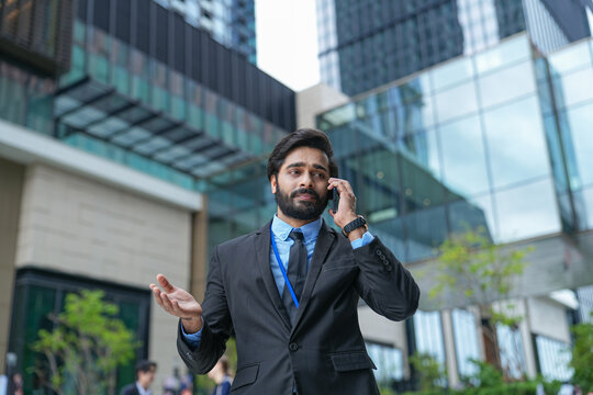A young Indian businessman concentrates on an important call outside the office tower in the green outdoor courtyard of a downtown business complex, his posture stays open and assertive