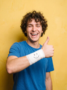 Smiling young man showing thumb up with bandaged wrist against yellow wall, cheerful and positive pose