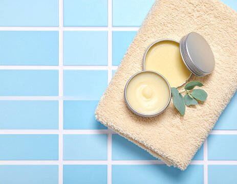 Opened cosmetic lip balm metal tin near beige bath towel on bathroom countertop with blue tiled floor, top view with copy space. Skincare and personal care concept