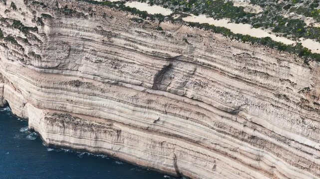 close detail of striated limestone cliff strata reveals layered rock bands descending toward deep blue mediterranean sea along malta coastline a thin green clifftop edge frames the geological texture