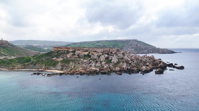 reddish sandstone headland juts into the mediterranean on maltas coast sea caves rock stacks ringing the point beside a sheltered cove sandy beach below green hillsides under grey sky