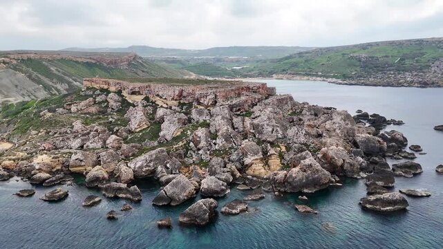 sea stacks boulders surround a rocky point on maltas west coast reddish layered cliffs rising above a curved bay with a sandy beach terraced green hillside under overcast sky