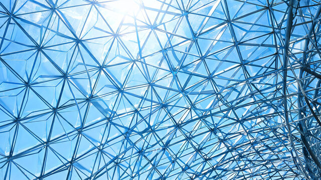 A modern geometric metal framework structure, with triangular beams forming a lattice, viewed against a bright blue sky with sunlight filtering through.