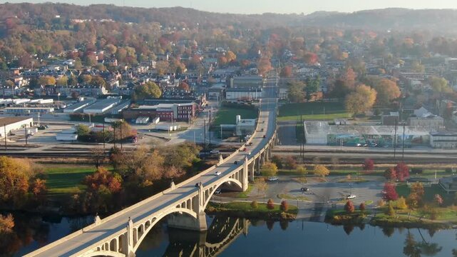 Aerial view of cars driving over a large arch bridge spanning a river, small town and autumn foliage in background.