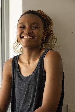 Adult African woman smiling, posing near vertical window at home, wearing dark gray ribbed tank top