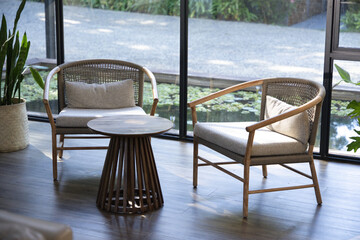 Fototapeta premium Pair of wooden lounge chairs are resting beside floor-to-ceiling glass windows with round table