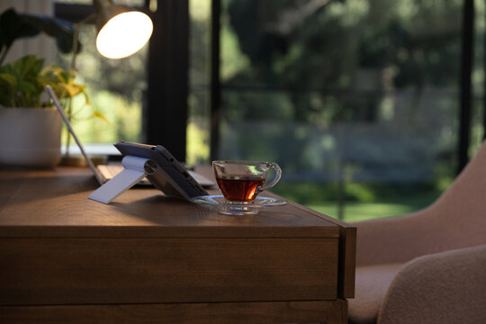 Clear glass teacup on saucer is sitting on wooden desk near large windows, with lit lamp