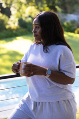 African woman standing on balcony in gray leaning on cable railing holding mug, smartwatch, pool © WavebreakMediaMicro