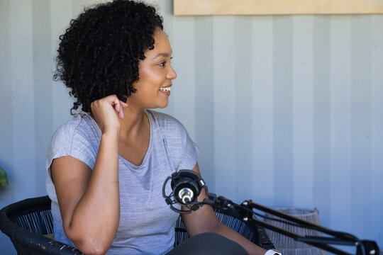 Adult African woman speaking into condenser microphone while sitting in woven chair at home studio