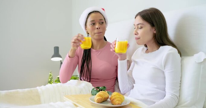 After toast gesture, diverse female friends tapping juice glasses and sharing croissants on bed
