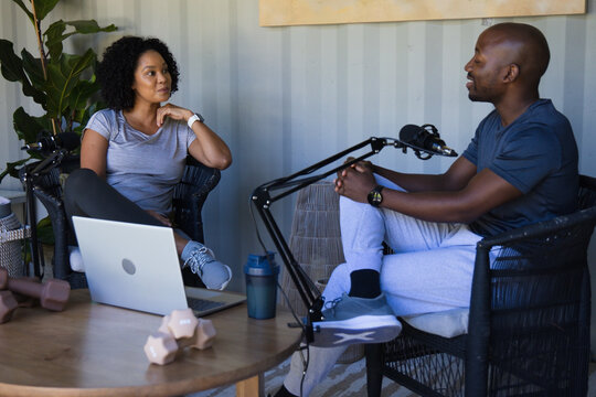 African American co-hosts sitting at table recording podcast with laptop, mics, wearing athleisure