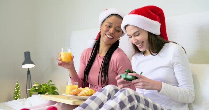 Diverse mother and daughter in Santa hats hugging, unwrapping gift, sipping juice from tray in bed