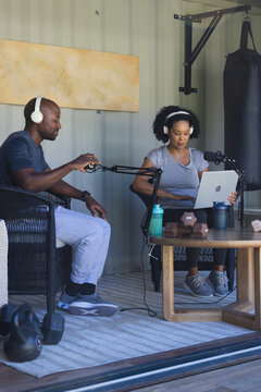 African American couple recording podcast in garage gym, sitting in athletic wear with mics, laptop
