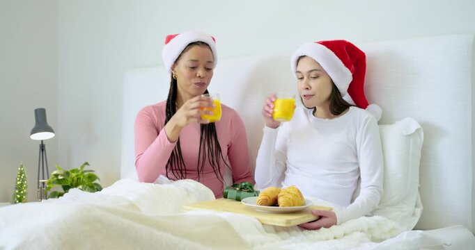 At dawn Diverse female friends in Santa hats toasting juice sharing breakfast croissants in bed