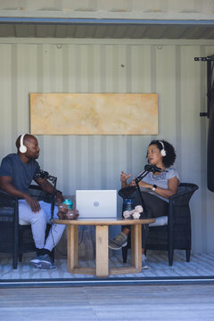 African American podcast co-hosts sitting in shipping container studio using microphones and laptop