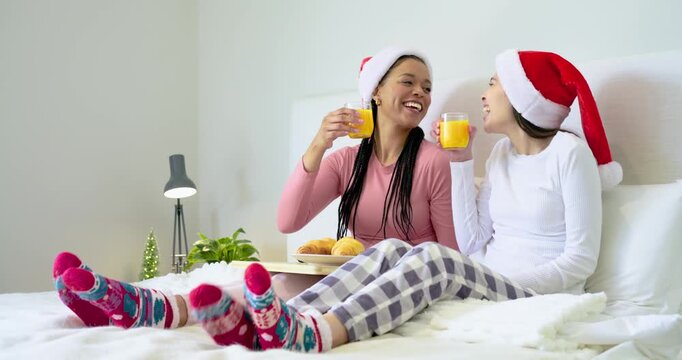 Smiling, African American and Asian women in Santa hats raising glasses, nibbling croissants on bed