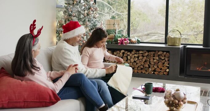 Child tearing wrap to reveal gift box while grandparents watching by Christmas tree copy space