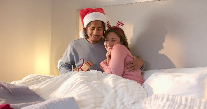 Diverse couple wearing Santa hat reacting to gift presentation laughing on bed, untying red ribbon