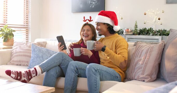 Upon smartphone alert, Diverse couple leaning on couch sipping mugs and sharing festive laughter