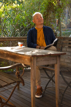 Weathered wooden table is holding open book and glass teacup on sheltered porch, sunlight filtering