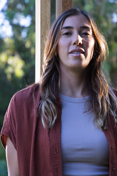Woman standing at doorframe on porch, wearing rust shirt and gray ribbed top amid green foliage
