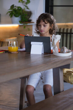 African American child sitting at dining table at home using tablet on stand, holding pencil