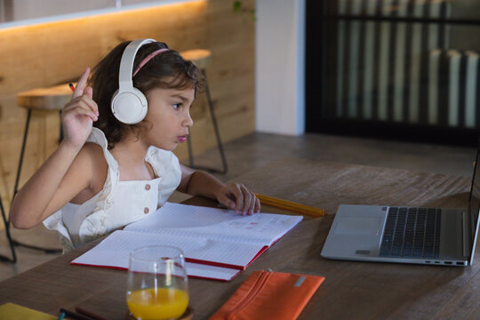 African American child girl sitting at table wearing white dress and headset raising hand to laptop