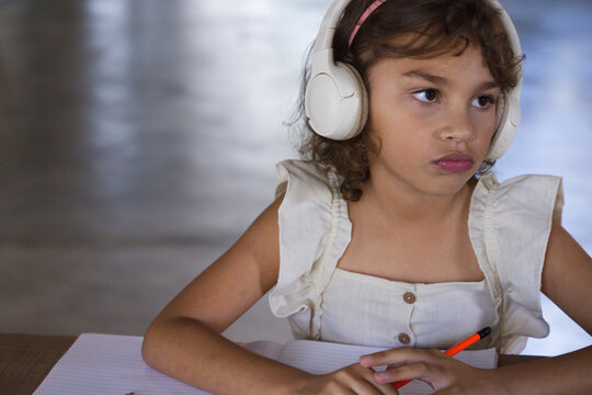 Girl sitting at table in hall wearing cream top and headphones, holding orange pencil and notebook