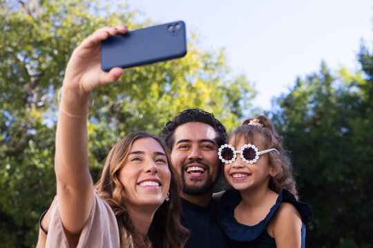 Diverse family taking selfie in park with black smartphone, child wearing white floral sunglasses