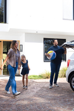 Family walking in driveway, child wearing navy dress sunglasses, father loading beach ball into SUV