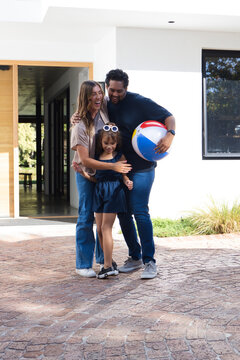 Family standing on driveway at home hugging, holding striped beach ball, wearing white sunglasses