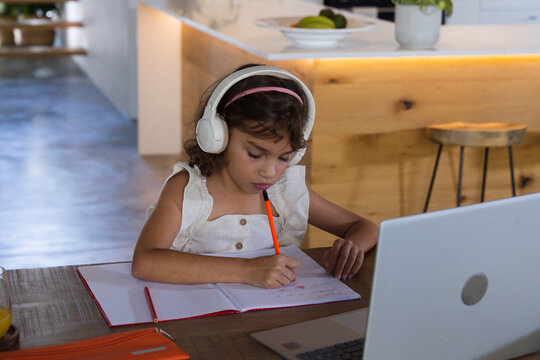 School-age girl wearing white sleeveless dress and headphones writing in red notebook at home table
