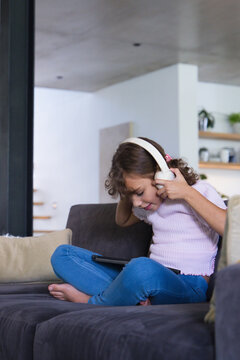 Female child sitting on gray sofa at home wearing pink top holding white headphones using tablet