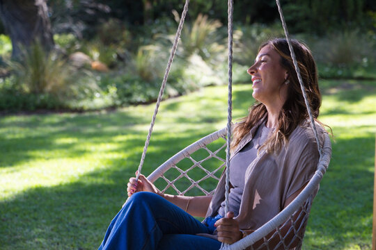 Adult woman sitting in white woven hanging chair in sunlit backyard holding rope, smiling