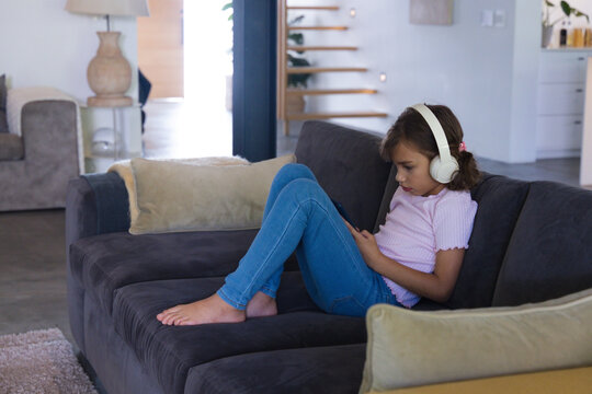 African American teen girl sitting on sofa holding phone, wearing headphones, pink top, copy space