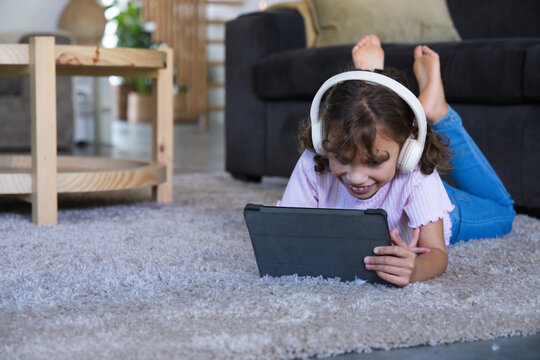 African American child lying on light gray rug using tablet, wearing white headphones, copy space